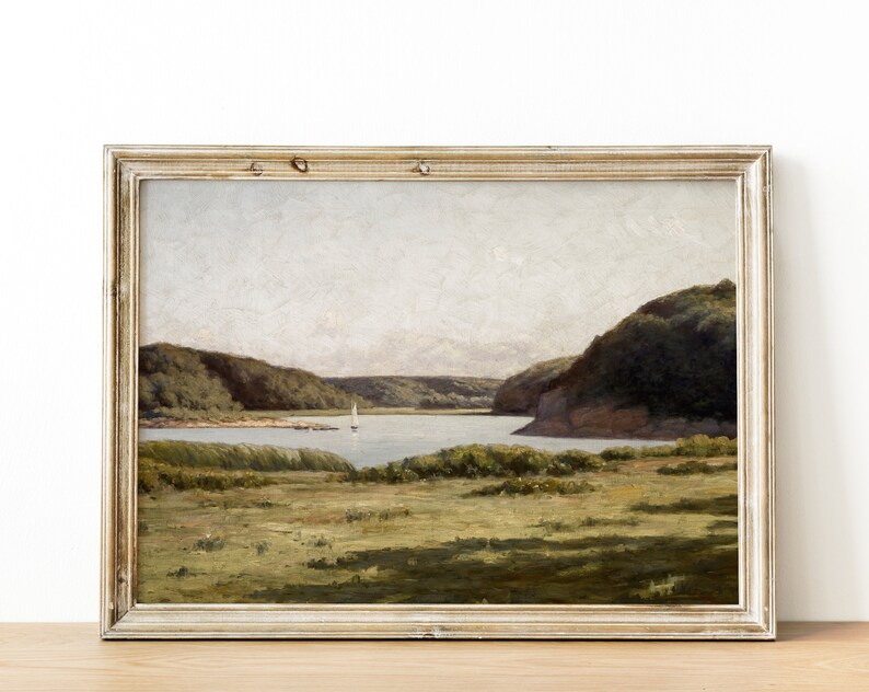 May include: A framed painting depicting a landscape scene with a lake, green hills, and a sailboat in the distance. The painting is set against a white wall.
