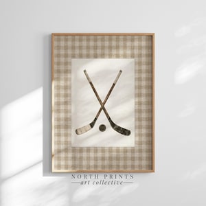 May include: Framed wall art featuring a vintage-style illustration of crossed hockey sticks and a puck. The artwork is set against a cream-colored background and framed with a light brown checkered pattern. The text "NORTH PRINTS - art collective -" is at the bottom.