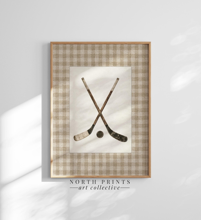 May include: Framed wall art featuring a vintage-style illustration of crossed hockey sticks and a puck. The artwork is set against a cream-colored background and framed with a light brown checkered pattern. The text "NORTH PRINTS - art collective -" is at the bottom.