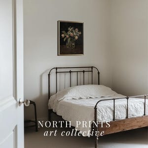 May include: A white bed with a black metal frame and a white bedspread. A framed painting of a bouquet of flowers in a vase is hanging on the wall behind the bed. The text "NORTH PRINTS art collective" is printed on the floor in front of the bed.