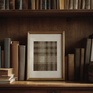 May include: A framed print with a brown and beige plaid pattern, displayed on a wooden shelf. The print is in a gold frame and surrounded by a collection of vintage books. The shelf is part of a dark wood bookcase.