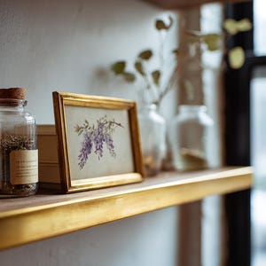 May include: A glass jar with a cork stopper and a label, filled with dried herbs, sits on a wooden shelf. Next to it is a framed print of purple wisteria flowers. The shelf has a gold trim.