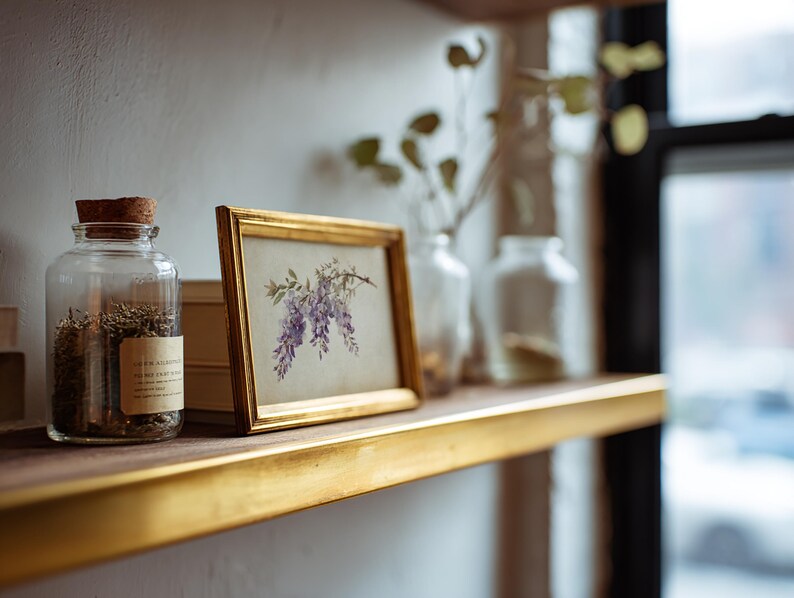 May include: A glass jar with a cork stopper and a label, filled with dried herbs, sits on a wooden shelf. Next to it is a framed print of purple wisteria flowers. The shelf has a gold trim.