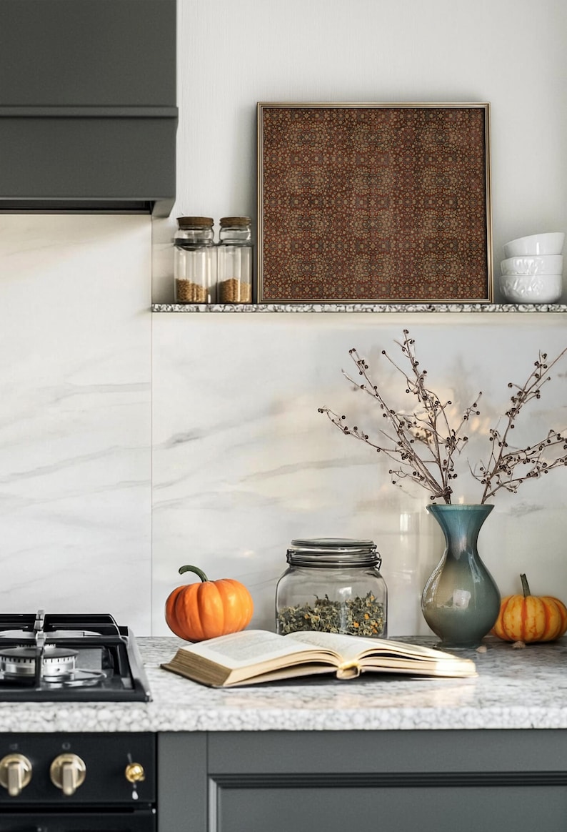 May include: A kitchen countertop scene featuring a gas hob, an open book, pumpkins, and a glass jar filled with herbs. A blue vase holds branches, and a framed artwork hangs above. The colour palette includes orange, brown, and white.