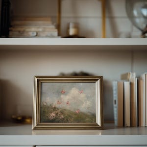 May include: A framed landscape painting with a gold-colored frame. The artwork depicts a field with pink flowers and butterflies against a cloudy sky. The painting is displayed on a white shelf with books.
