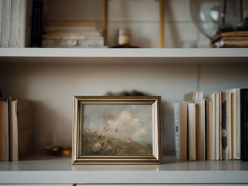 May include: A framed landscape painting with a gold-colored frame. The artwork depicts a field with pink flowers and butterflies against a cloudy sky. The painting is displayed on a white shelf with books.