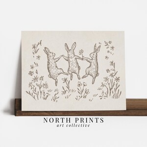 May include: A black and white line drawing of three rabbits dancing in a field of flowers. The rabbits are holding hands and appear to be having fun. The drawing is on a light brown background.