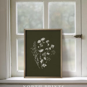 May include: A framed botanical print featuring white floral silhouettes on a dark green background. The gold frame surrounds the artwork, which is displayed on a white windowsill. The text "NORTH PRINTS art collective" is at the bottom.