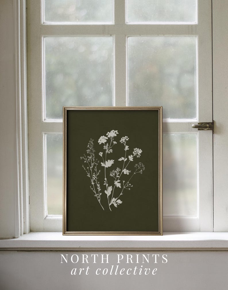 May include: A framed botanical print featuring white floral silhouettes on a dark green background. The gold frame surrounds the artwork, which is displayed on a white windowsill. The text "NORTH PRINTS art collective" is at the bottom.