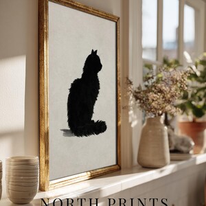 Peut inclure: Impression d'art encadr&eacute;e repr&eacute;sentant une silhouette de chat noir sur fond neutre, dans un cadre dor&eacute;. L'&oelig;uvre fait partie de la collection "art collective". L'impression est expos&eacute;e sur une &eacute;tag&egrave;re blanche pr&egrave;s d'une fen&ecirc;tre.