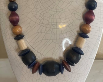 May include: A wooden bead necklace with a variety of colors and shapes, including brown, red, blue, and white. The necklace is made of natural wood and has a rustic look.