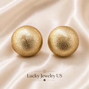 Peut inclure: Deux boucles d'oreilles à clous dorées et texturées. Les boucles d'oreilles sont rondes et ont une finition mate. Elles sont présentées sur un tissu satiné crème. Le texte "Lucky Jewelry US" est en bas de l'image.