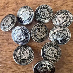 May include: A collection of silver-coloured buttons with a raised design featuring a Native American chief in a headdress. The buttons are circular and have the words "United States of America" and the year "1776" around the edge.