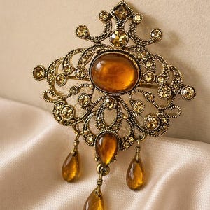 May include: An ornate, vintage-style brooch in antique gold. The brooch features an oval amber-colored center stone, smaller amber-colored stones, and three amber teardrop dangles. The design includes intricate filigree and scrollwork.