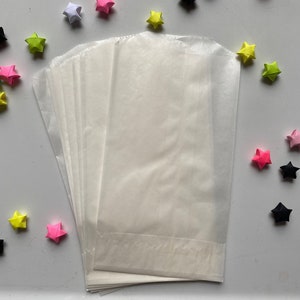 Puede incluir: Un montón de bolsas de papel blanco translúcido, posiblemente para empacar artículos pequeños. Alrededor, estrellas de origami coloridas en tonos rosa, amarillo, verde y negro, creando un arreglo decorativo.