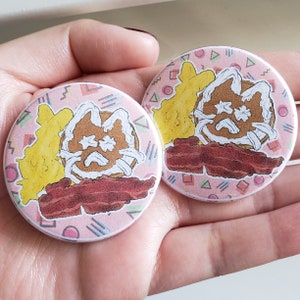 May include: Two round pink and white buttons with a cartoon illustration of a pancake with a face, bacon, and scrambled eggs. The buttons have a 90s style geometric pattern.