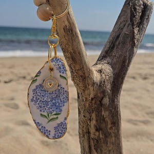 May include: A keychain featuring a gold-toned metal clasp adorned with four round wooden beads. Attached is a white oyster shell painted with blue hydrangea flowers and green leaves, accented by a small circular charm with a gold heart emblem.
