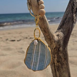 May include: A keychain featuring a gold-toned metal ring adorned with five smooth, light-colored wooden beads. A gold lobster clasp connects to a circular ring holding a flat, shell-shaped pendant with a blue and white striped pattern and gold trim.