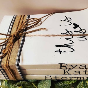 以下が含まれることがあります： 黒い文字で書かれた3冊の白い本の積み重ね。一番上の本には、ハートのマークと共に「This is us」という文字が筆記体で書かれている。2番目の本には「Ryan」という名前が書かれている。3番目の本には「Kate」と「Storm」という名前が書かれており、足跡のマークが付いている。本は、黒と白のチェック柄のリボンと麻紐で包まれている。