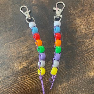 Puede incluir: Dos llaveros de cuentas de arcoíris con cierres plateados y cuerda morada. Las cuentas son blancas, azul claro, azul oscuro, verdes, amarillas, naranjas y rojas.