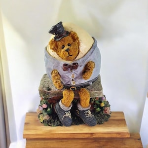 May include: A whimsical figurine of a teddy bear emerging from an egg, wearing a top hat, bow tie, and a button-down shirt. The bear is seated on a stone base with floral accents, and wearing shoes. The figurine is on a wooden surface.