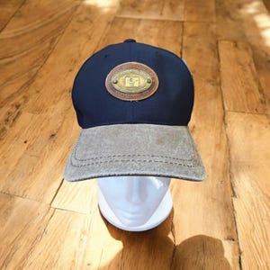 May include: A navy blue baseball cap with a gray brim. The cap features a leather patch with the text "Cleveland Cavaliers" and a basketball graphic. The hat is displayed on a white mannequin head.