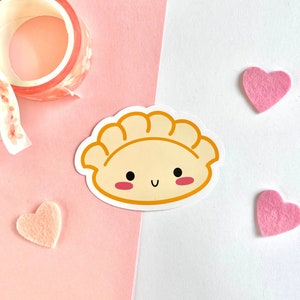 May include: A cute dumpling sticker with a smiling face and rosy cheeks. The sticker is on a white background with pink felt hearts and a roll of floral washi tape.