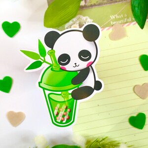Op de afbeelding: Een groene en witte panda-sticker die een groene boba-theebeker vasthoudt. De panda heeft een schattige uitdrukking op zijn gezicht en houdt de beker vast met zijn poten. De boba-theebeker bevat bruine boba-parels.