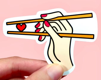 May include: A hand with red nail polish holds a pair of chopsticks with a red heart between them.