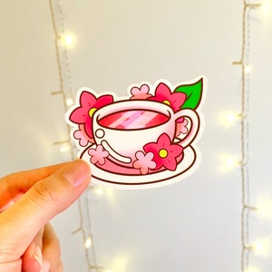 May include: A white teacup with pink flowers and a green leaf on a pink saucer. The teacup is filled with pink liquid.