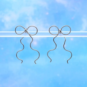 May include: Two silver-colored bow-shaped earrings with a wavy, elongated design. The earrings are suspended against a bright blue sky background, creating a minimalist and elegant aesthetic. The bows are attached to a clear, horizontal bar.