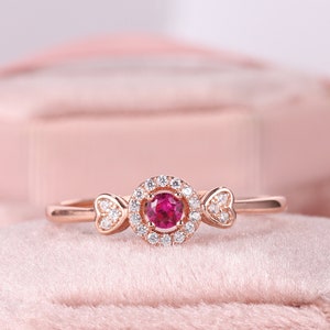 Puede incluir: Anillo de oro rosa con una gema roja redonda central, rodeada de piedras blancas más pequeñas. Detalles en forma de corazón flanquean la piedra central. El anillo se muestra sobre un fondo rosa suave. Diseño delicado y elegante.