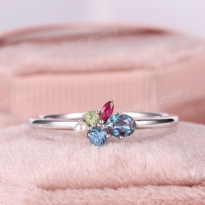 May include: A delicate silver ring with a cluster of colorful gemstones. The ring features a small pearl, a green gemstone, a red gemstone, and two blue gemstones.