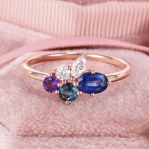 May include: A rose gold ring with a cluster of gemstones. The ring features a large oval blue sapphire, two smaller blue sapphires, a round white diamond, two marquise-shaped white diamonds, and a round amethyst.