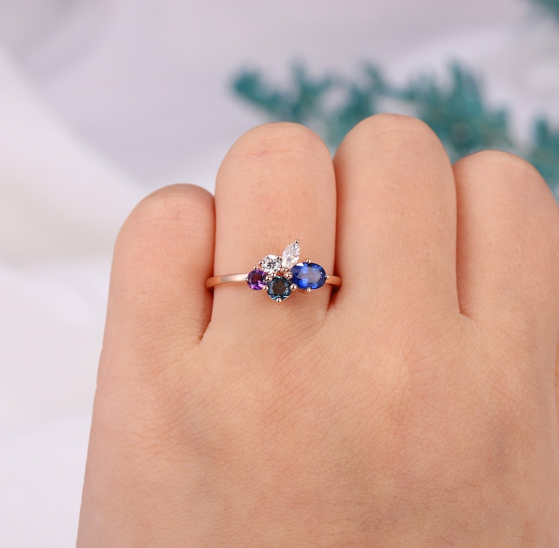 May include: A rose gold ring with a cluster of colorful gemstones. The ring features a large oval blue sapphire, a smaller round blue sapphire, a round purple amethyst, and two round white diamonds.