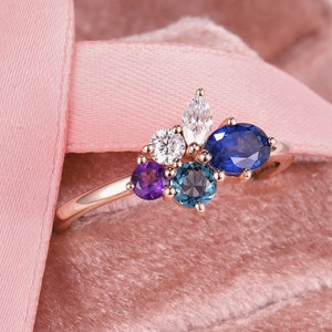May include: A rose gold ring with a cluster of colorful gemstones. The ring features a large oval sapphire, a smaller round sapphire, a pear-shaped diamond, a round diamond, and two amethyst stones.
