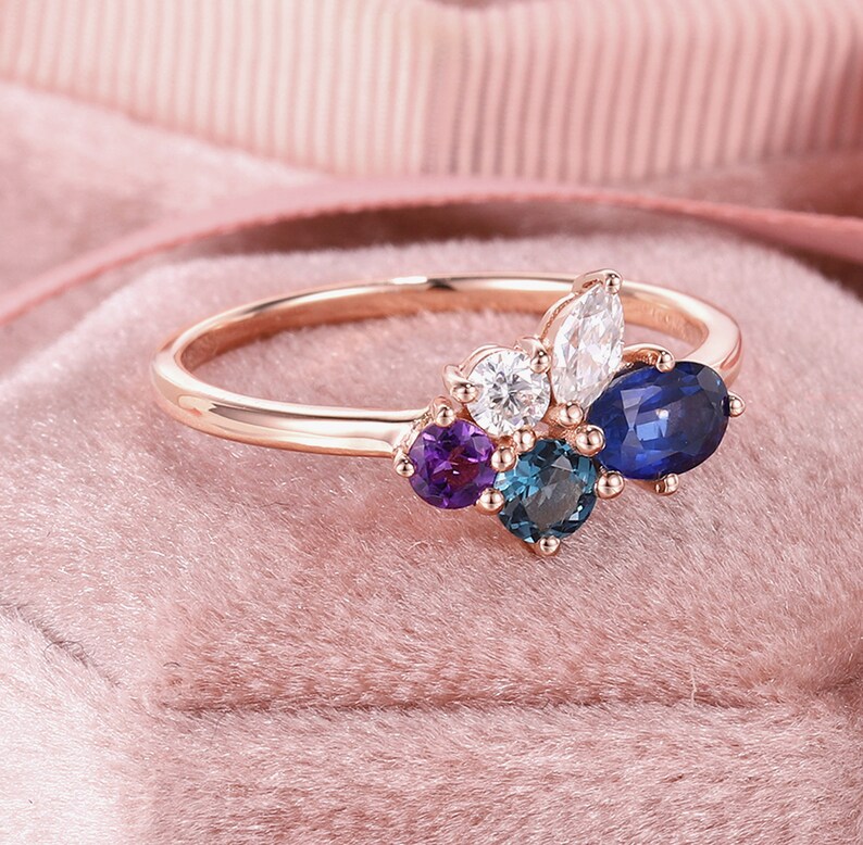 May include: A rose gold ring with a cluster of gemstones. The ring features a large oval sapphire, a smaller round sapphire, a pear-shaped diamond, a round diamond, and a round amethyst.