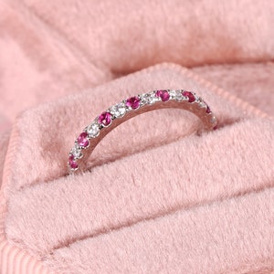 Op de afbeelding: Een delicate zilveren bandring met afwisselende roze en witte edelstenen. De ring ligt op een roze fluwelen achtergrond.