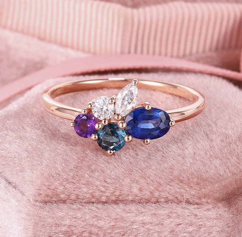 May include: A rose gold ring with a cluster of gemstones. The ring features a large oval blue sapphire, two smaller blue sapphires, a round white diamond, two marquise-shaped white diamonds, and a round amethyst.