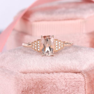May include: A rose gold ring with a large, rectangular, light pink morganite gemstone surrounded by a row of smaller, round, white diamonds.