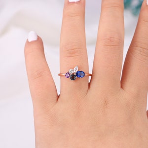 May include: A rose gold ring with a cluster of oval-shaped gemstones. The ring features a blue sapphire, a purple amethyst, a white diamond, and a smaller blue sapphire.