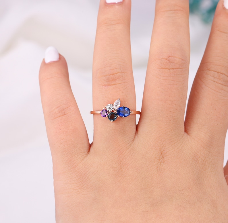 May include: A rose gold ring with a cluster of oval-shaped gemstones. The ring features a blue sapphire, a purple amethyst, a white diamond, and a smaller blue sapphire.