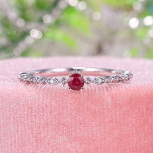 Op de afbeelding: Een delicate zilveren ring met een rode robijn-edelsteen omringd door kleinere diamanten. De ring heeft een delicaat bloemmotief.