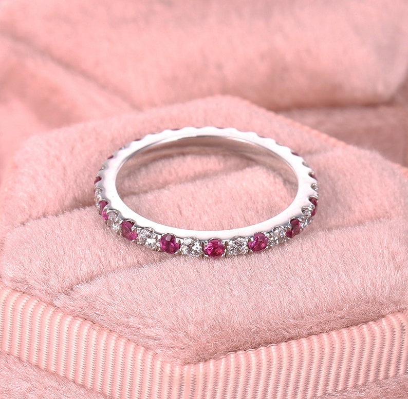Op de afbeelding: Een delicate eeuwigheidsring met een rij kleine, ronde, rode robijnen en witte diamanten gezet in een witte metalen band.