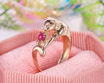 Puede incluir: Un anillo de oro con un diseño de pantera detallado y una piedra preciosa roja. La pantera sostiene la piedra preciosa en sus patas.