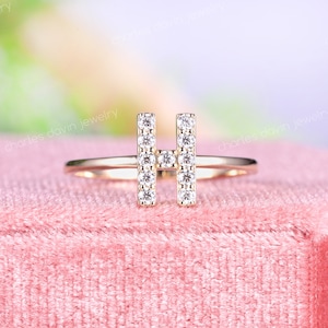 以下が含まれることがあります： ピンクのベルベット生地の上に置かれた、小さなダイヤモンドでできたHの文字がついた金の指輪です。