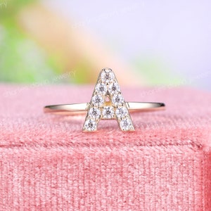 Puede incluir: Un anillo de oro con una letra A hecha de pequeños diamantes. El anillo está sobre una superficie de terciopelo rosa.