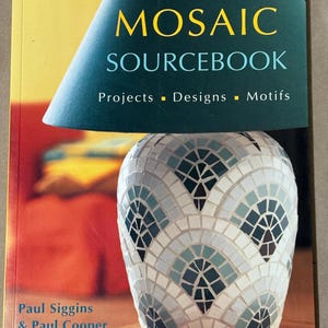 Op de afbeelding: The Mosaic Sourcebook van Paul Siggins en Paul Cooper. De omslag toont een mozaïekvaas met een waaiervormig ontwerp in tinten wit, grijs en blauw. De titel is in geel en blauw, met de woorden "Projects, Designs, Motifs".