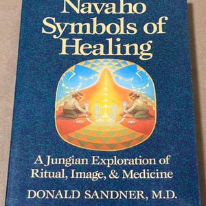 Puede incluir: Un libro titulado "Navaho Symbols of Healing" con una cubierta azul y texto blanco. La portada presenta una ilustración de dos figuras en un diseño circular. También es visible el nombre del autor, Donald Sandner, M.D.