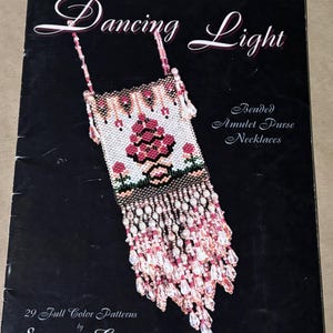 May include: A book cover titled Dancing Light Beaded Amulet Purse Necklaces by Suzanne Cooper featuring 29 Full Color Patterns. The cover displays a rectangular beaded pouch with a pink floral design and long, dangling pink and clear crystal beads.